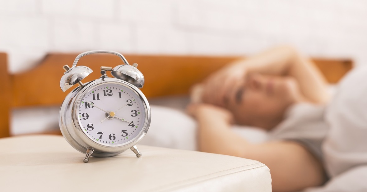Imagen de un reloj despertador y una mujer mayor que sufre un trastorno del sueño tumbada en la cama sin poder dormir