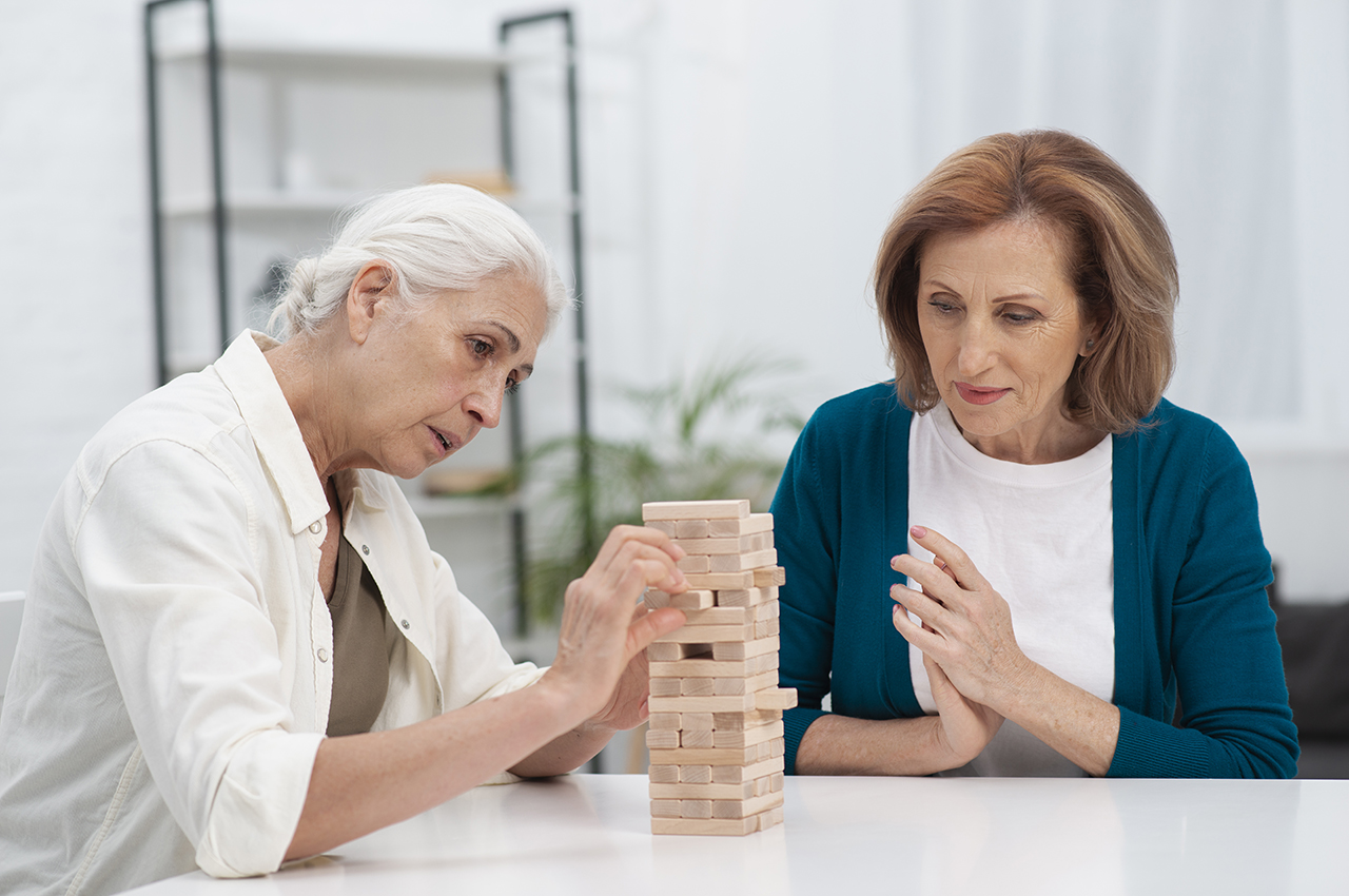 Dos mujeres, una más mayor que la otra, juegan al jenga. Este juego se basa en pequeñas piezas que forman una construcción que no debe caerse mientras, de forma estratégica, calculan cómo puede seguir manteniéndose la torre cada vez más alta a pesar de las piezas restantes.