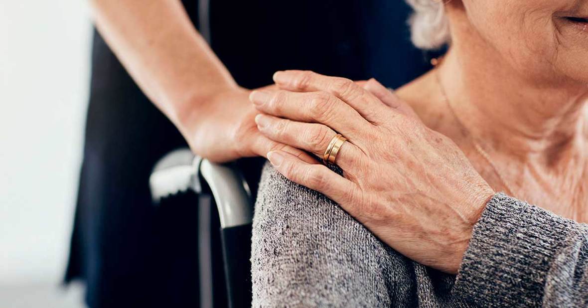 Mujer mayor dependiente con escaras o úlceras por presión sentada en una silla de ruedas y con la mano de su cuidadora tocándole el hombro