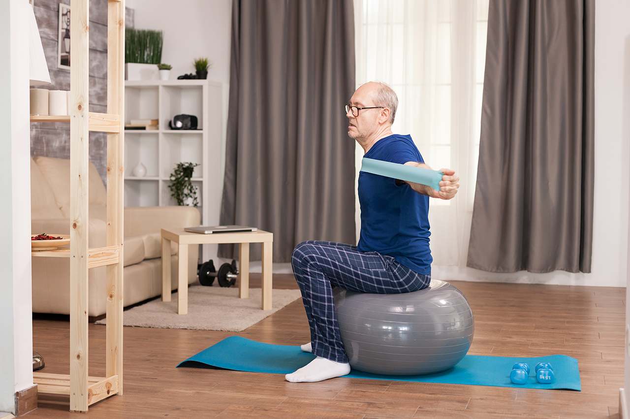Un hombre practica pilates en su salón. Está sentado en una bola de pilates mientras ejercita los brazos estirando una banda elástica. A su vez, tiene otros materiales cerca, como unas mancuernas de 1,5 kgs y una esterilla.