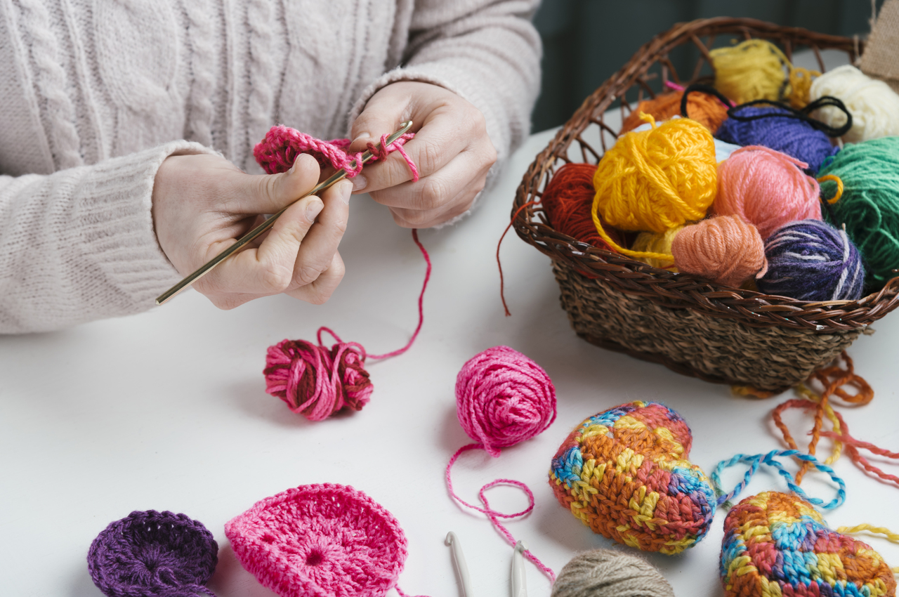 Una mujer haciendo crochet. A su vez, tiene al lado una cesta llena de ovillos de diversos colores y distintas manualidades ya terminadas o en proceso.