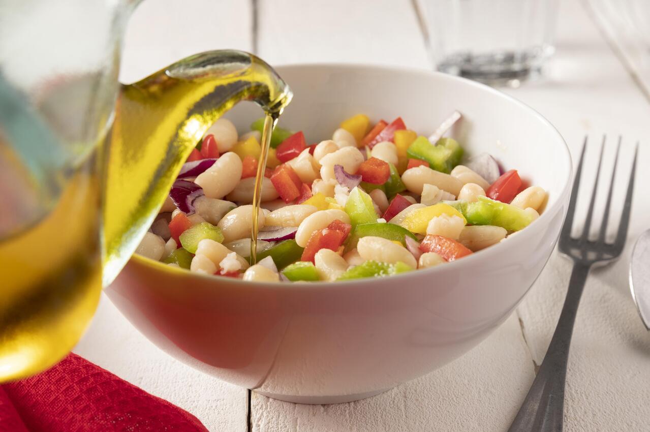 Una ensalada de legumbres, en la que predominan las judías blancas, tomate, pimiento y garbanzos. Están aderezándola con un chorrito de aceite.