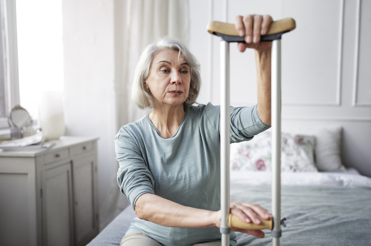 Una mujer mayor intenta levantarse de la cama ayudada por una muleta.