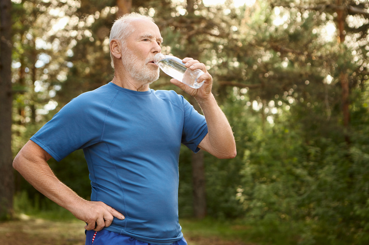 Un señor mayor, vestido con ropa deportiva, bebe un trago de agua desde una botella en plena naturaleza.