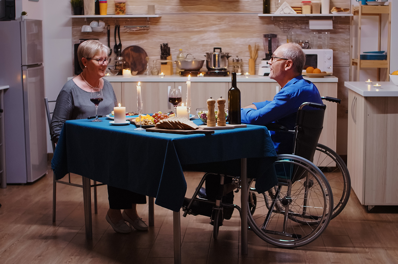 Una pareja de personas mayores, él en silla de ruedas, cena en una amplia cocina.