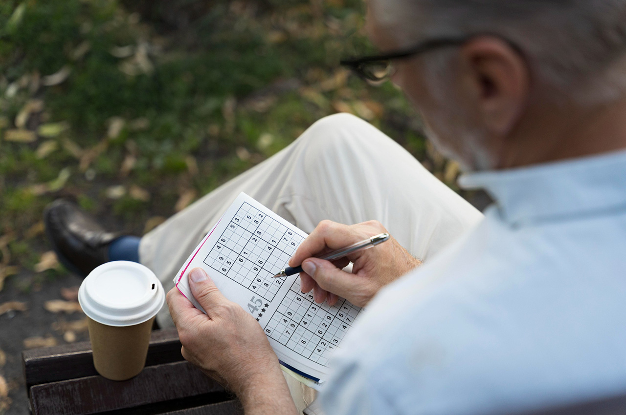 Un hombre mayor haciendo un sudoku sentado en un banco