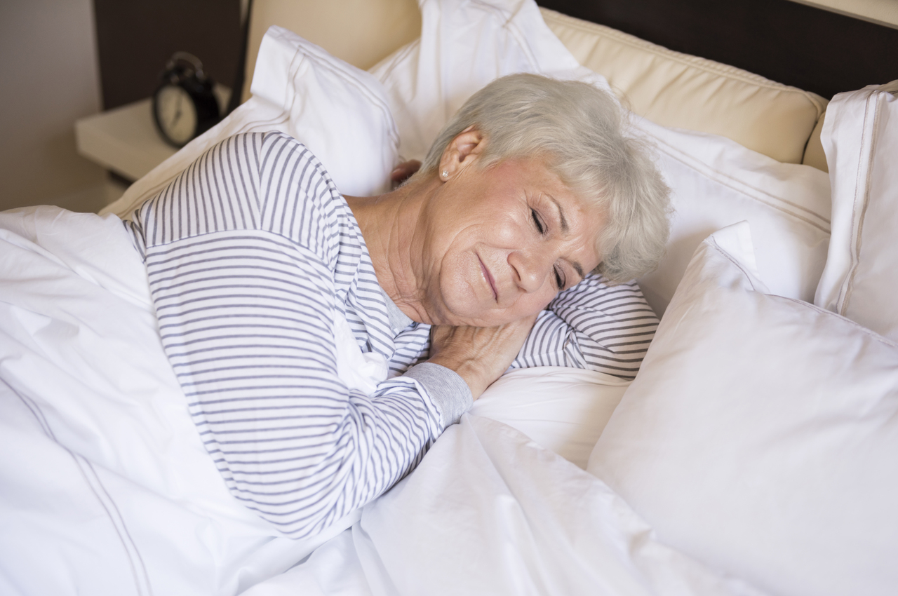 Una mujer mayor medita apaciblemente en su cama, tumbada de costado.