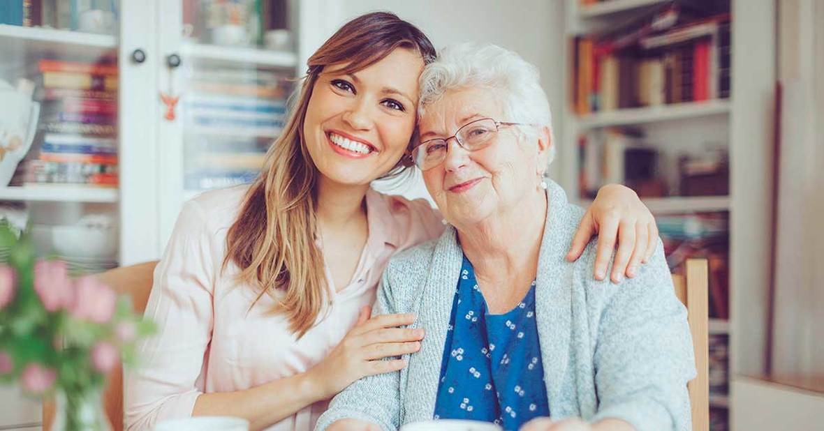 Cuidadora de personas mayores rodeando con sus brazos los hombros de una anciana mientras las dos sonríen