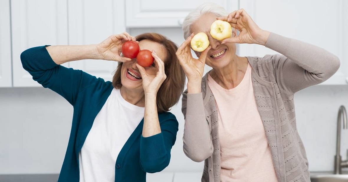 Una mujer mayor con dos tomates y otra mujer mayor con dos pimientos simulando que son sus ojos, mientras se divierten juntas en una cocina