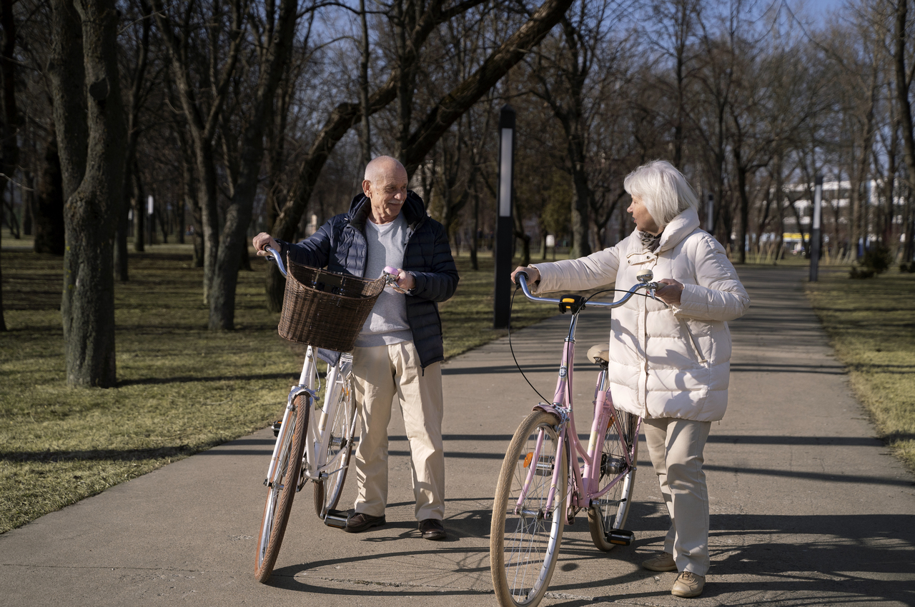 Dos personas mayores caminando, llevando cada uno una bicicleta, en un parque.