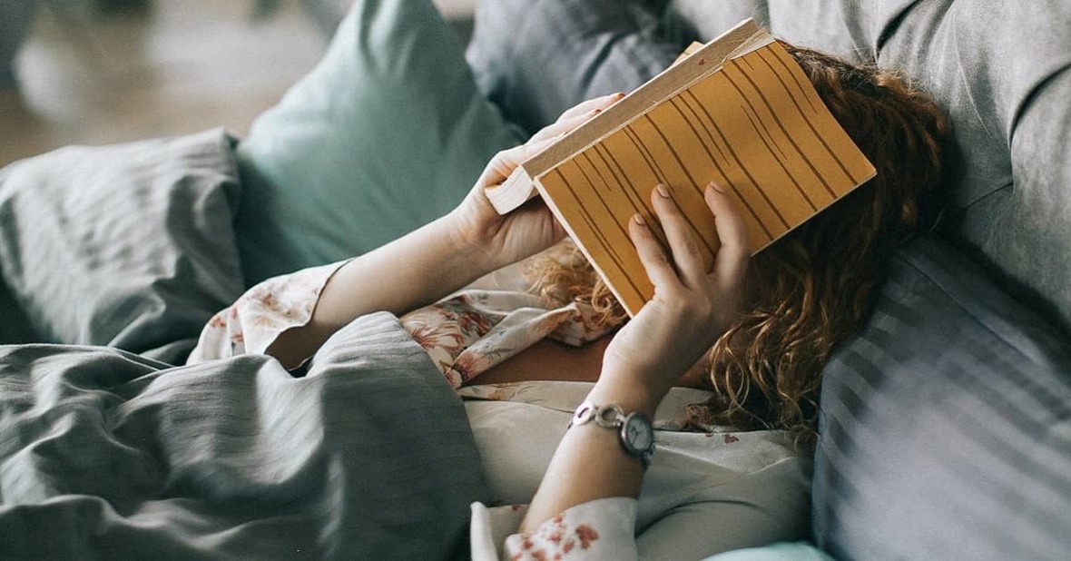 Una mujer leyendo en la cama, apoyando el libro sobre su cara, porque no tiene fuerza en los brazos debido a la fatiga crónica