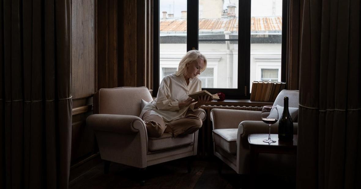 Mujer mayor leyendo un libro sentada en una butaca de su casa al lado de la ventana