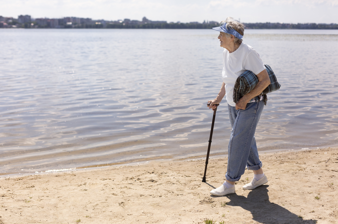 Una señora mayor camina ayudada de un bastón mientras visita una playa.