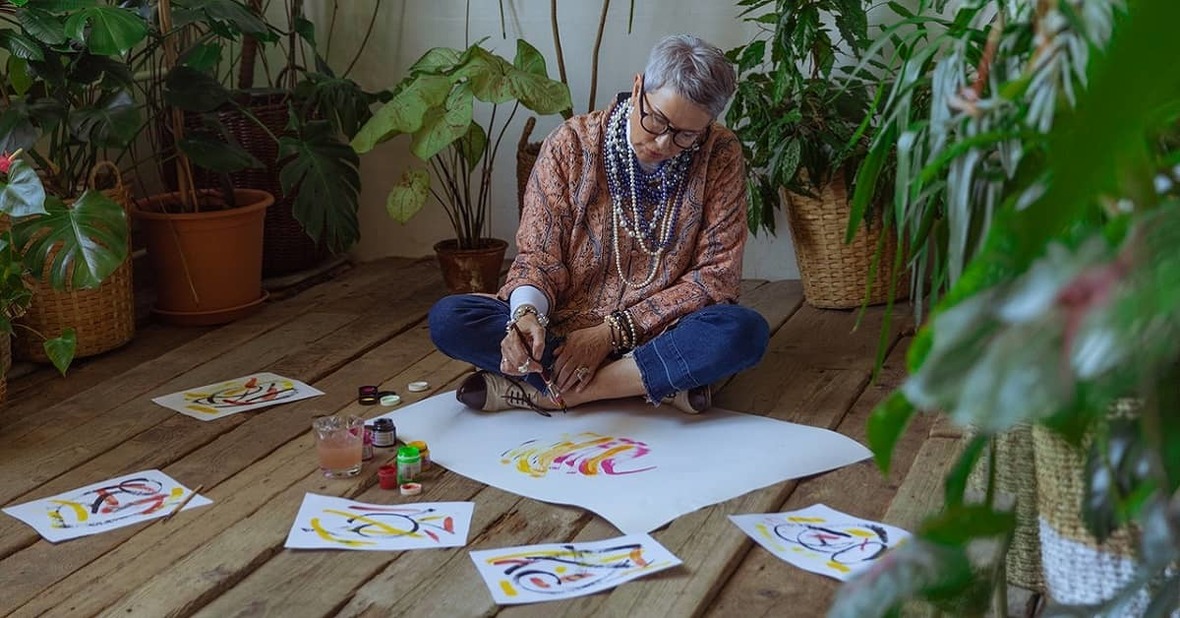 Mujer mayor pintando con témperas sentada en el suelo y rodeada de papeles con dibujos ya terminados