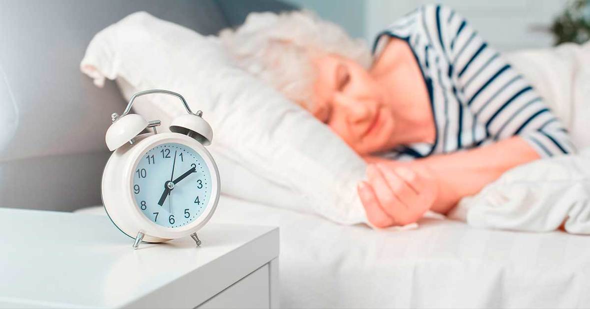 Mujer mayor con mucho sueño tumbada en la cama con un despertador en la mesilla de noche