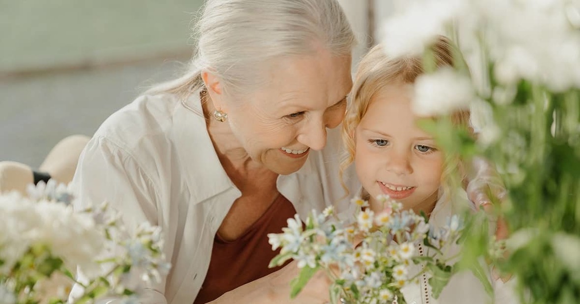Una niña mirando un jarrón con flores, mientras escucha lo que su abuela le está enseñando