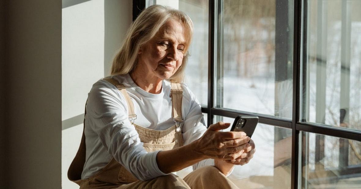Mujer mayor sentada frente a una ventana mirando su teléfono móvil con tristeza