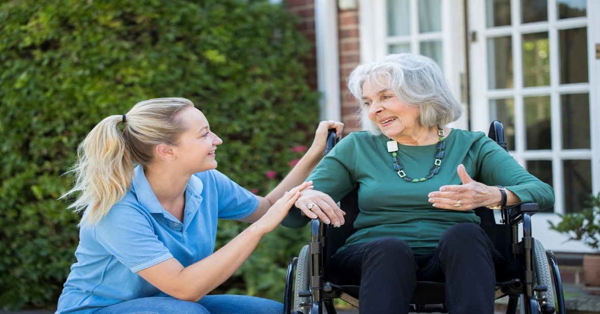 Joven cuidadora de rodillas sonriendo a una anciana con demencia senil en silla de ruedas