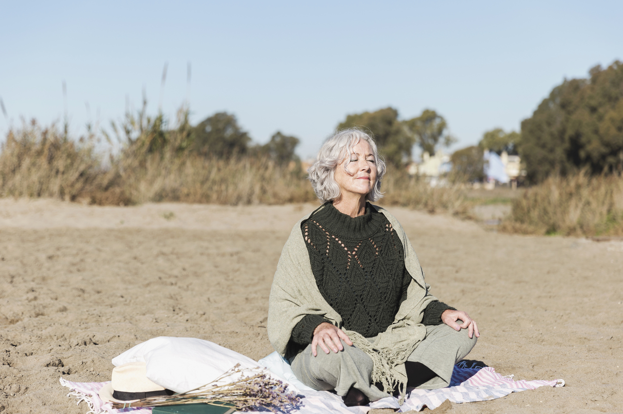 Una mujer mayor practica respiración guiada en la playa.