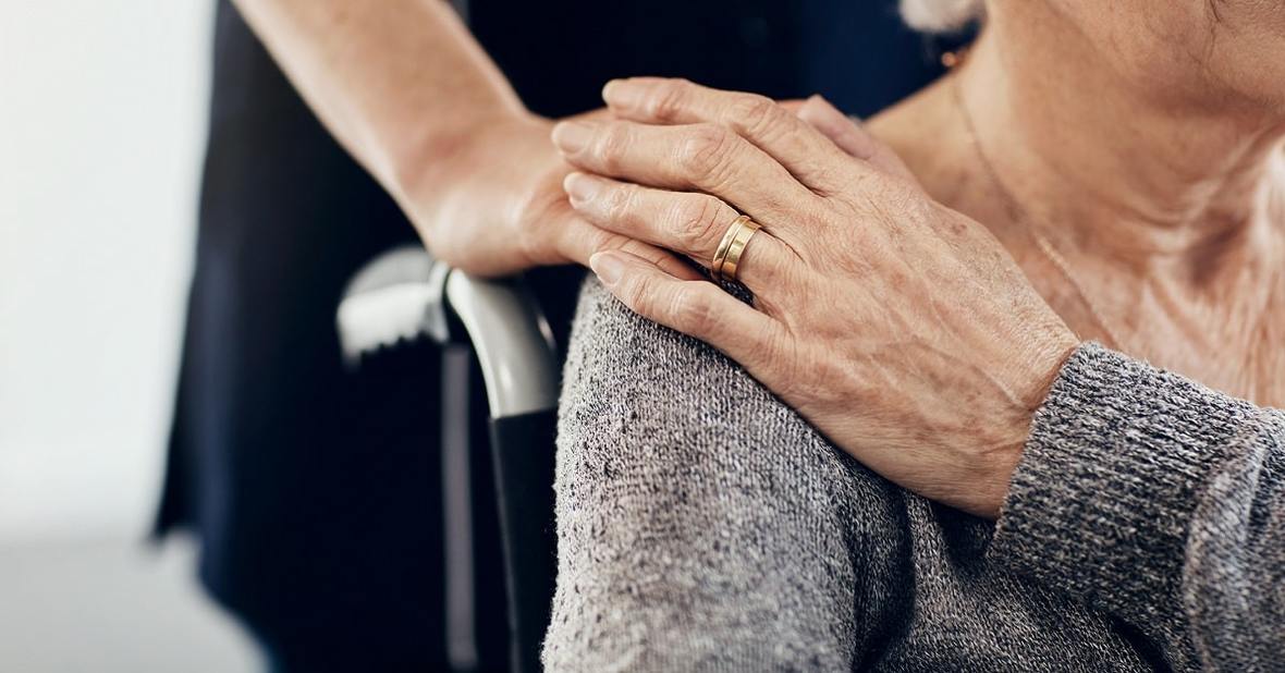Joven apoyando la mano en el hombro de una mujer mayor en silla de ruedas