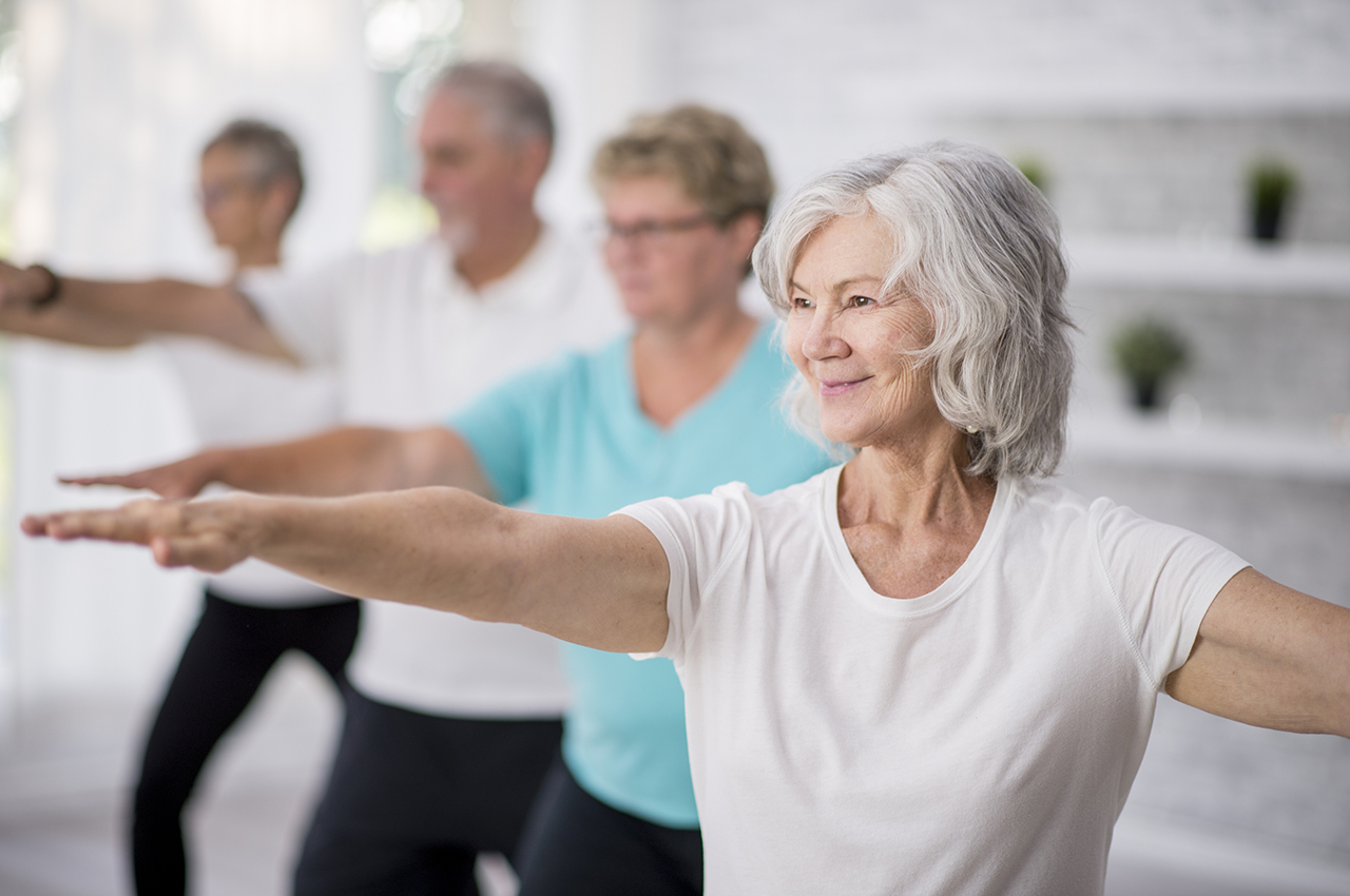 Un grupo de cuatro adultos mayores (dos hombres al fondo, dos mujeres más próximas a la cámara) realizan pilates. Ahora mismo, están haciendo un ejercicio para fortalecer brazos, con ellos extendidos delante y detrás del tronco.