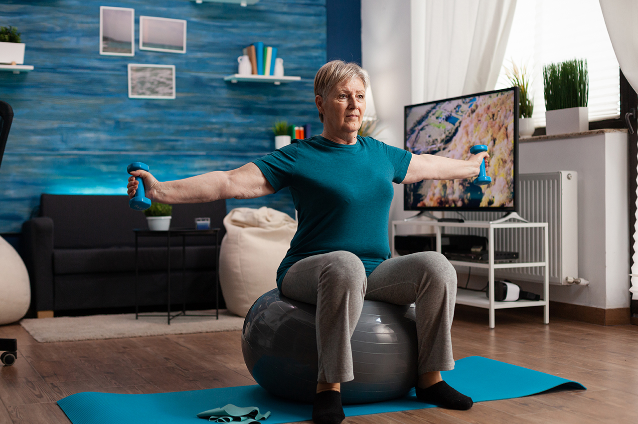 Una mujer mayor hace ejercicios en casa. En este momento, está fortaleciendo los brazos utilizando unas pesas ligeras mientras se sienta en un balón suizo (típico al practicar pilates)