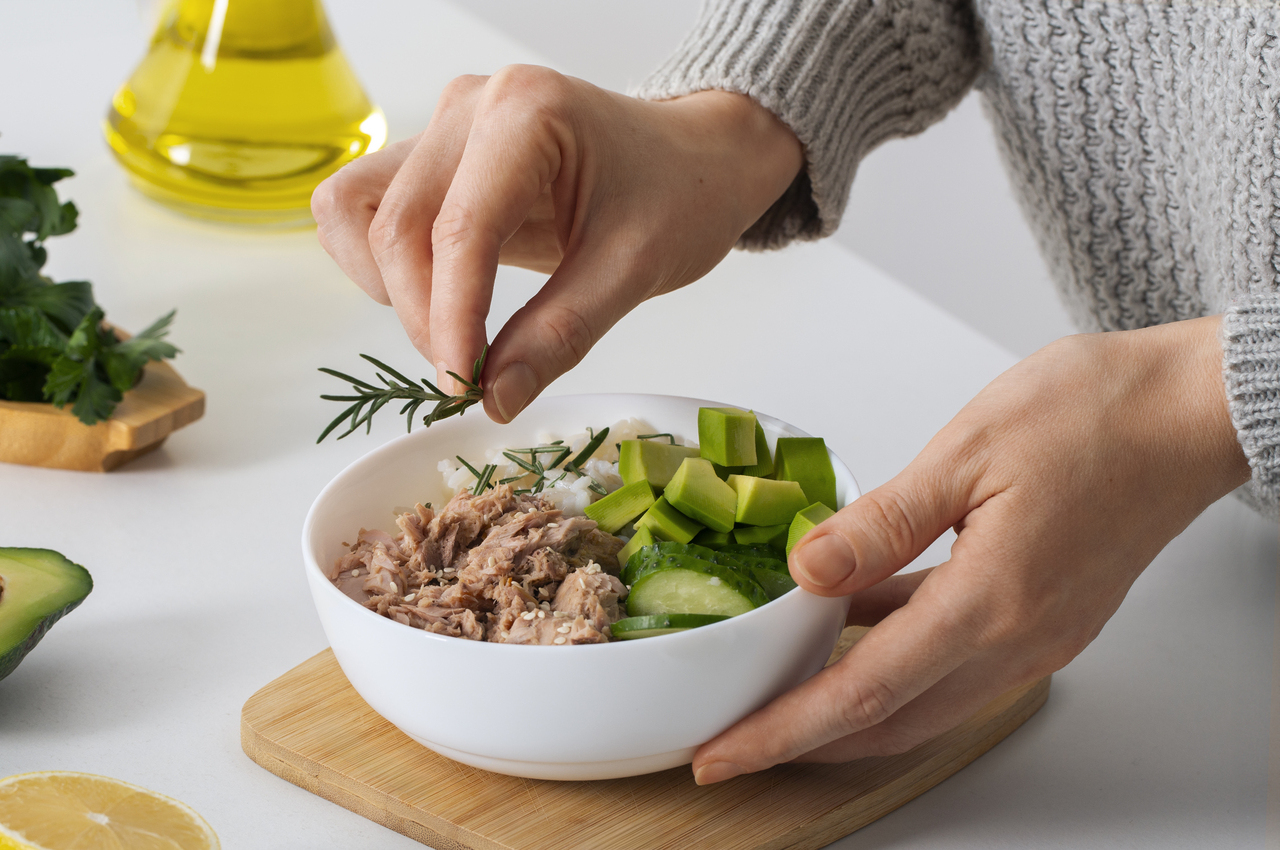 Una persona adereza con romero un bol con atún, arroz, pepino y aguacate.